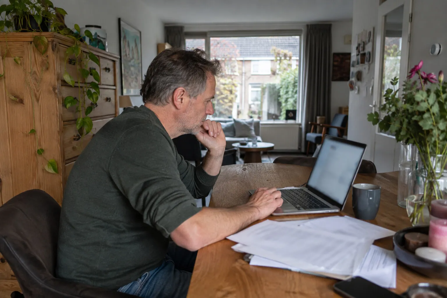 Man aan de eettafel zoekt iets op zijn laptop naast een stapel uitgeprinte documenten