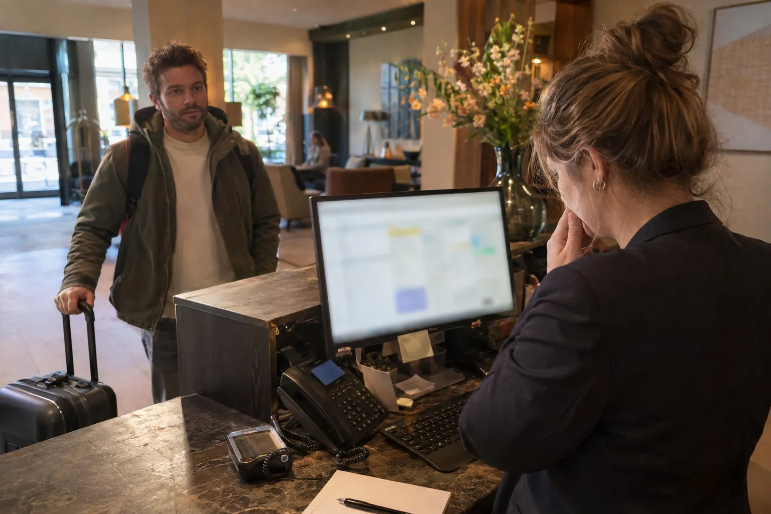 Receptionist van een hotel kijkt bezorgd naar zijn scherm terwijl een gast met koffer wacht