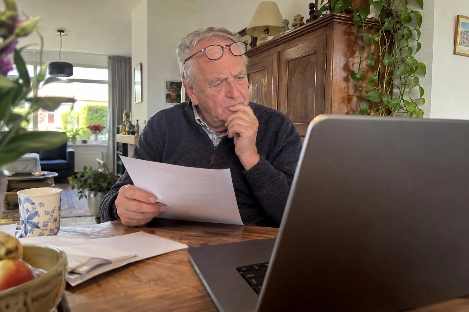 Oudere man aan de eettafel met een geprinte rekening en een laptop, twijfelend