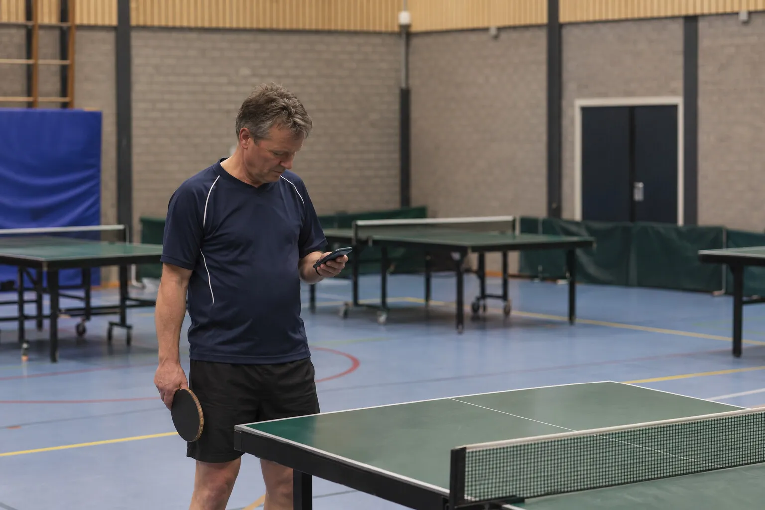 Tafeltennisser met batje in een sporthal kijkt naar zijn telefoon